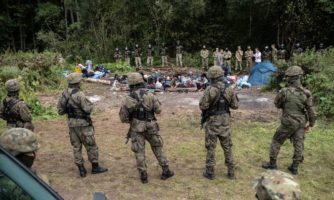 Grupa żołnierzy stojąca naprzeciw grupy zablokowanych migrantów.