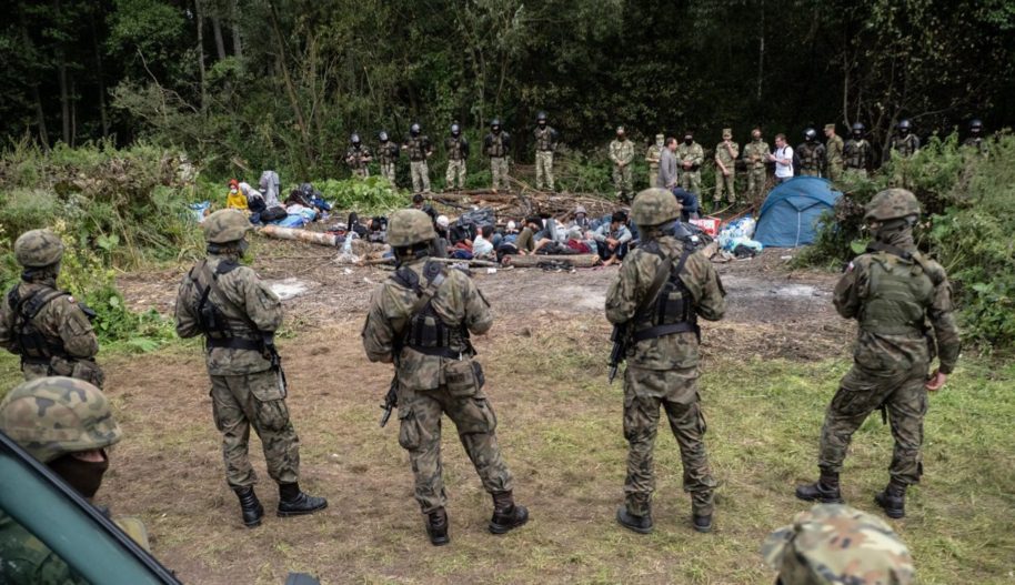 Grupa żołnierzy stojąca naprzeciw grupy zablokowanych migrantów.