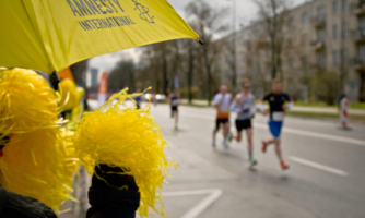 Zdjęcie z punktu kibicowania przy maratonie. Na pierwszym tle żółte pompony i parasol z logo Amnesty International. W tle biegną uczestnicy maratonu.