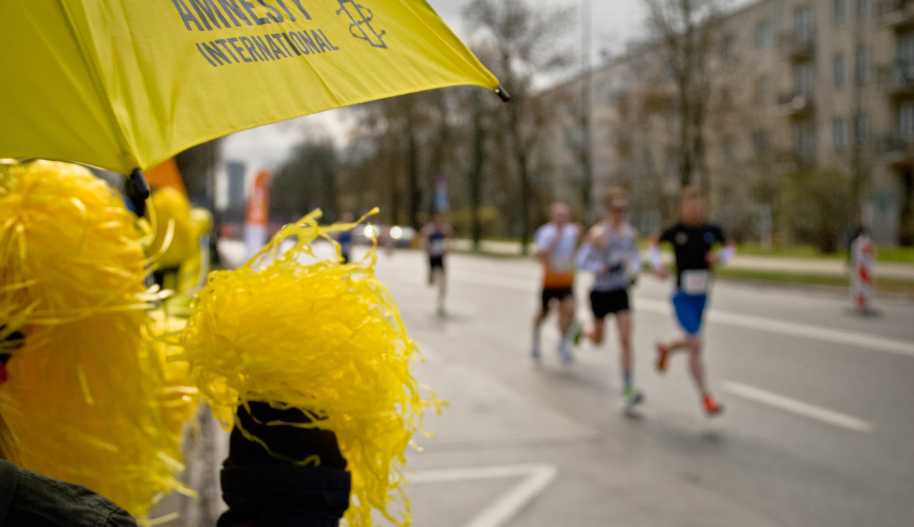 Zdjęcie z punktu kibicowania przy maratonie. Na pierwszym tle żółte pompony i parasol z logo Amnesty International. W tle biegną uczestnicy maratonu.